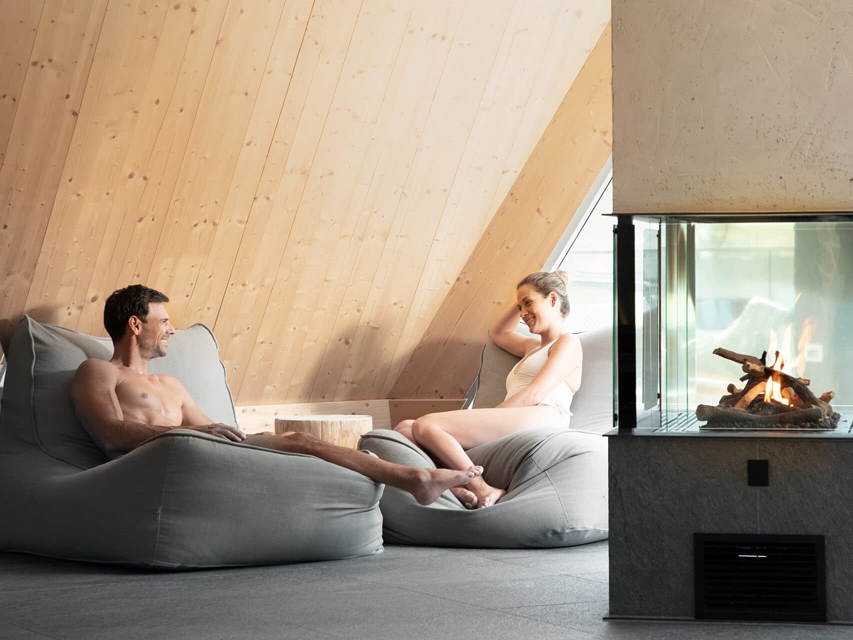 Ein Mann und eine Frau entspannen auf Sitzsäcken in einem gemütlichen Raum mit Holzwänden. Ein Feuer in einem modernen Kamin sorgt für Wärme.