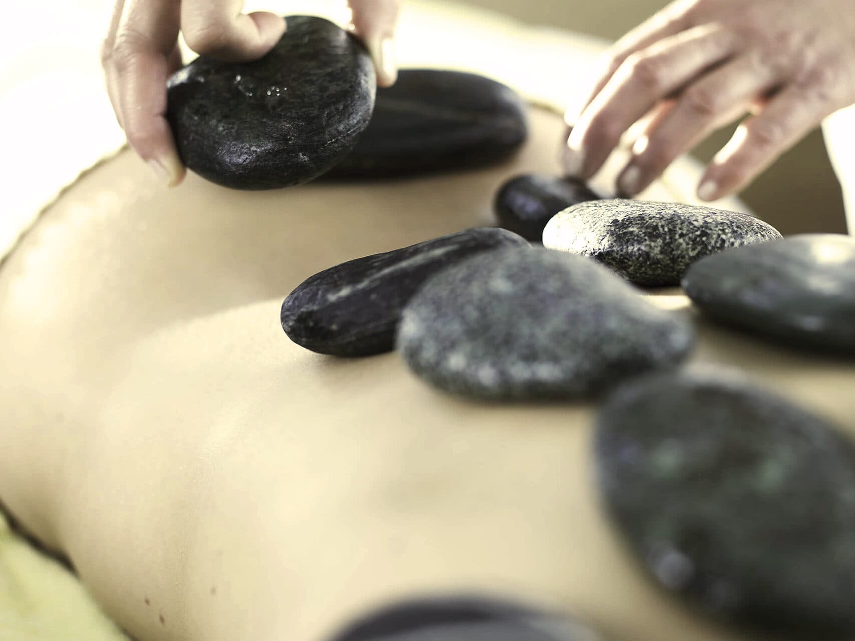 Eine Person erhält eine Hot-Stone-Massage. Dabei werden mehrere glatte, dunkle Steine auf ihren Rücken gelegt. Die Hände eines Therapeuten richten die Steine aus.