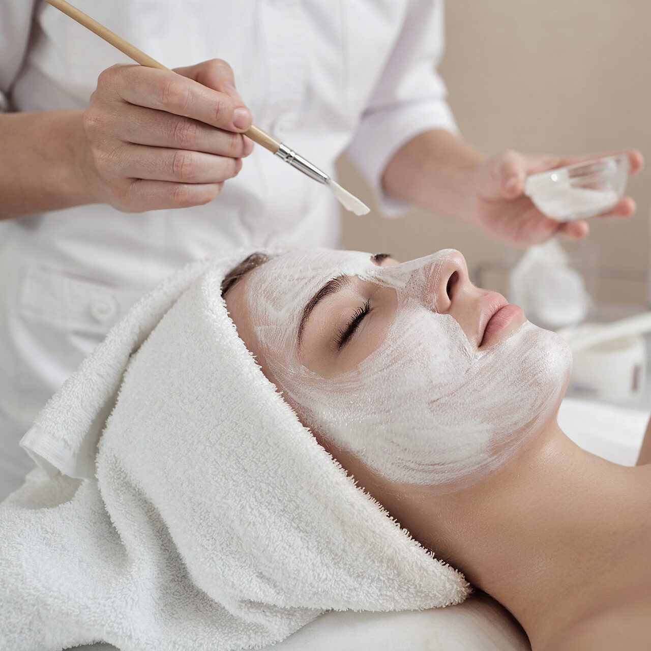 Person erhält eine Gesichtsbehandlung mit einer weißen Maske, die von einem Fachmann mit einem Pinsel aufgetragen wird. Die Person liegt mit einem Handtuch um den Kopf.