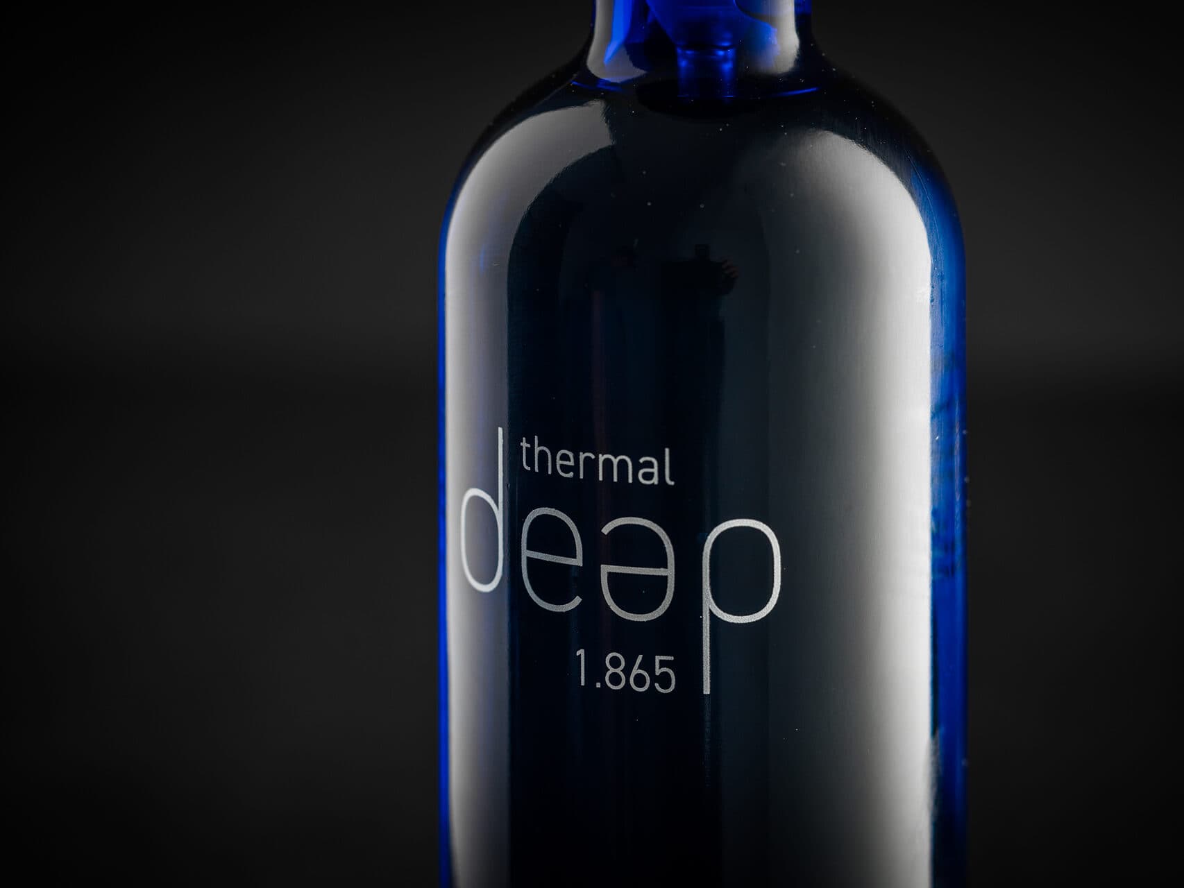 Nahaufnahme einer blauen Glasflasche mit dem weißen Text „thermal deep 1.865“. Der dunkle Hintergrund hebt die reflektierende Oberfläche der Flasche hervor.