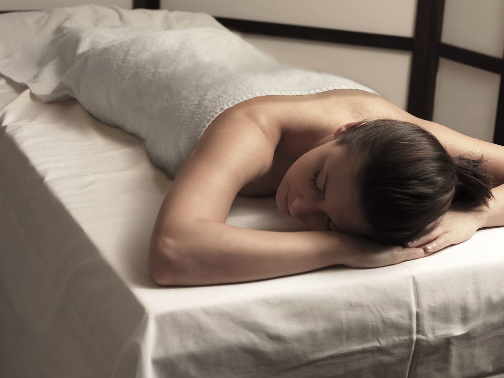 Person, die mit dem Gesicht nach unten auf einem Massagetisch liegt, bedeckt mit einem weißen Handtuch in einem beruhigenden, sanft beleuchteten Raum.