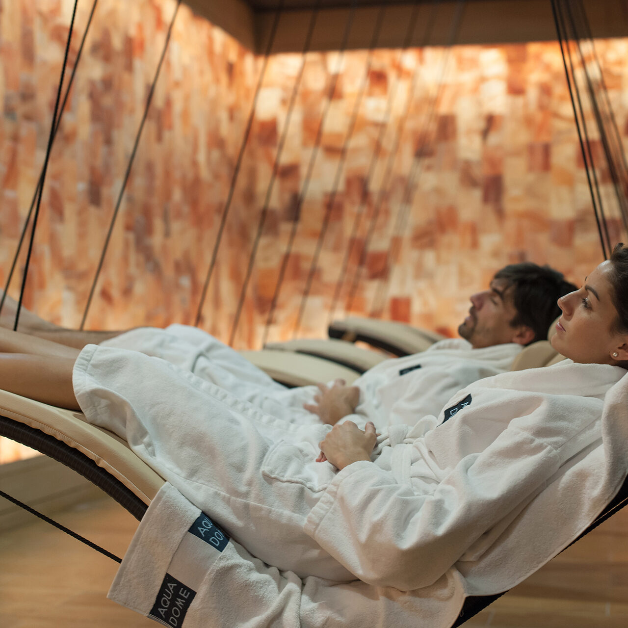 Zwei Menschen in weißen Bademänteln entspannen auf Hängeliegen in einem Wellnessraum mit Salzsteinwand und Holzboden.