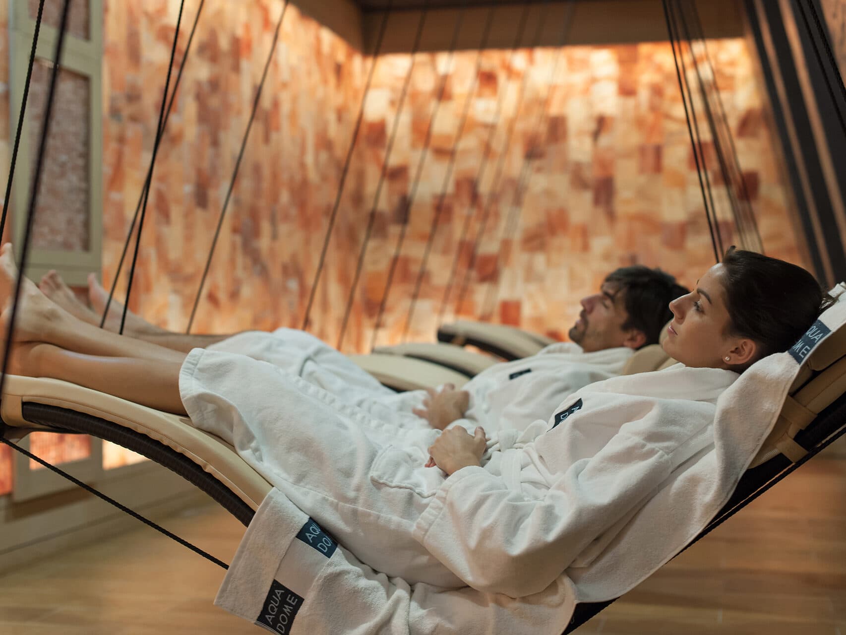 Zwei Menschen in weißen Bademänteln entspannen auf Hängeliegen in einem Wellnessraum mit Salzziegelwänden.