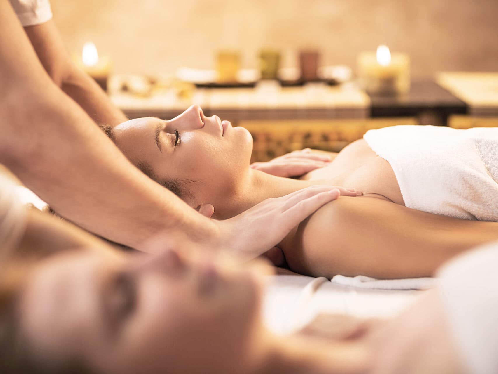 Eine Frau erhält eine Nacken- und Schultermassage in einer entspannenden Spa-Umgebung mit Kerzen und sanfter Beleuchtung im Hintergrund.
