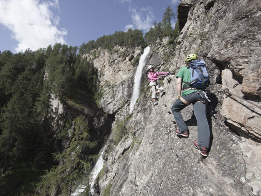 Двое скалолазов в касках и снаряжении поднимаются по крутой скале рядом с водопадом, в окружении деревьев и ясного неба.