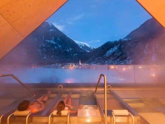 Two people unwind in a heated outdoor spa pool, gazing at the snowy mountain landscape at dusk, under a warm-lit triangular roof.