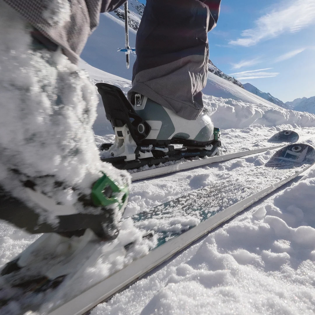 Nahaufnahme der Stiefel und Skier eines Skifahrers auf Schnee, mit schneebedeckten Bergen und einem klaren blauen Himmel im Hintergrund.
