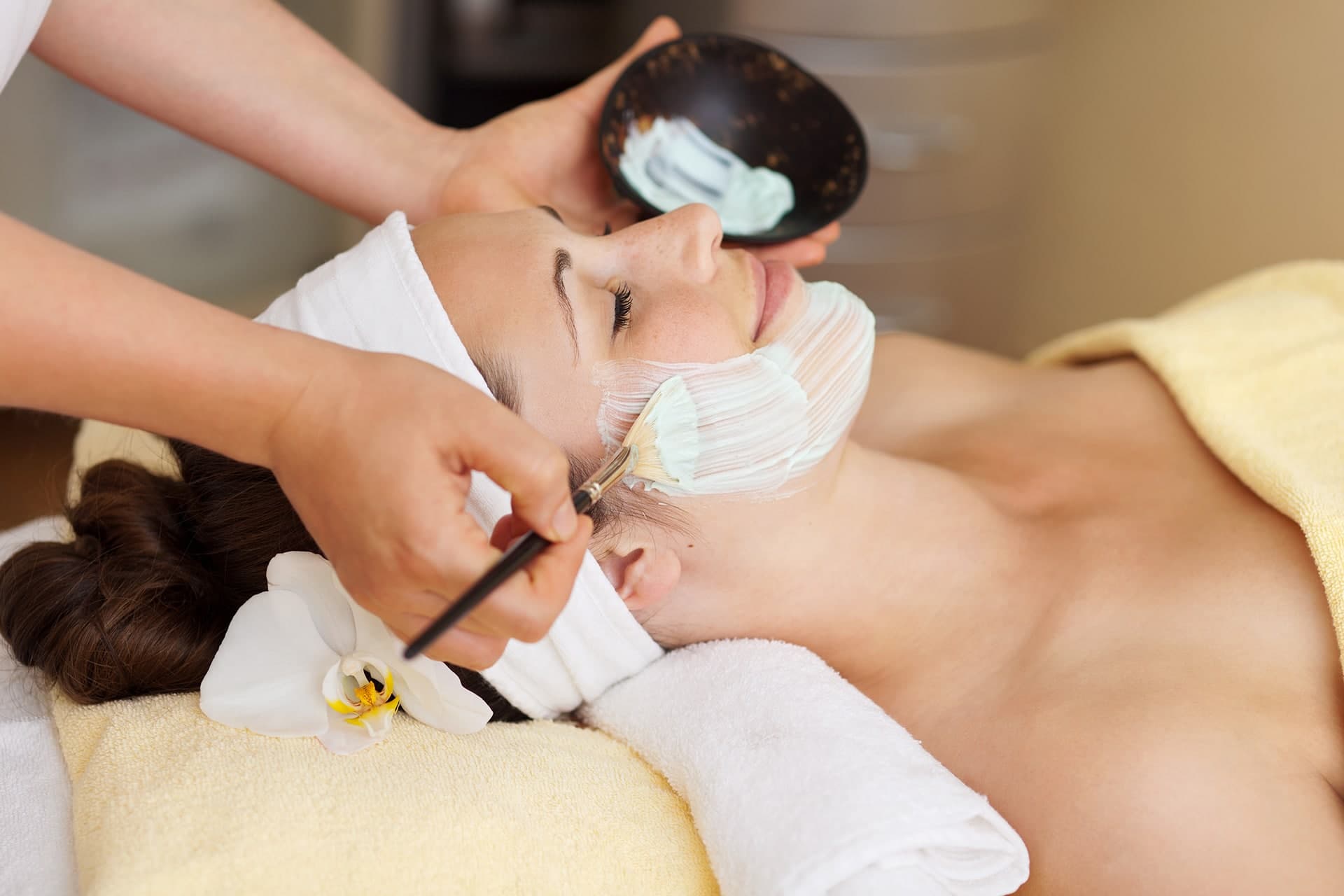 Person receiving a facial treatment with a green mask applied using a brush, lying on a spa table with a yellow towel and white flowers nearby.