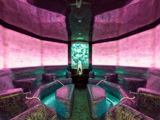 A tranquil spa seating area boasts a reflective ceiling, enhanced by soothing pink and green lighting, with tile benches arranged around an inviting central water feature.