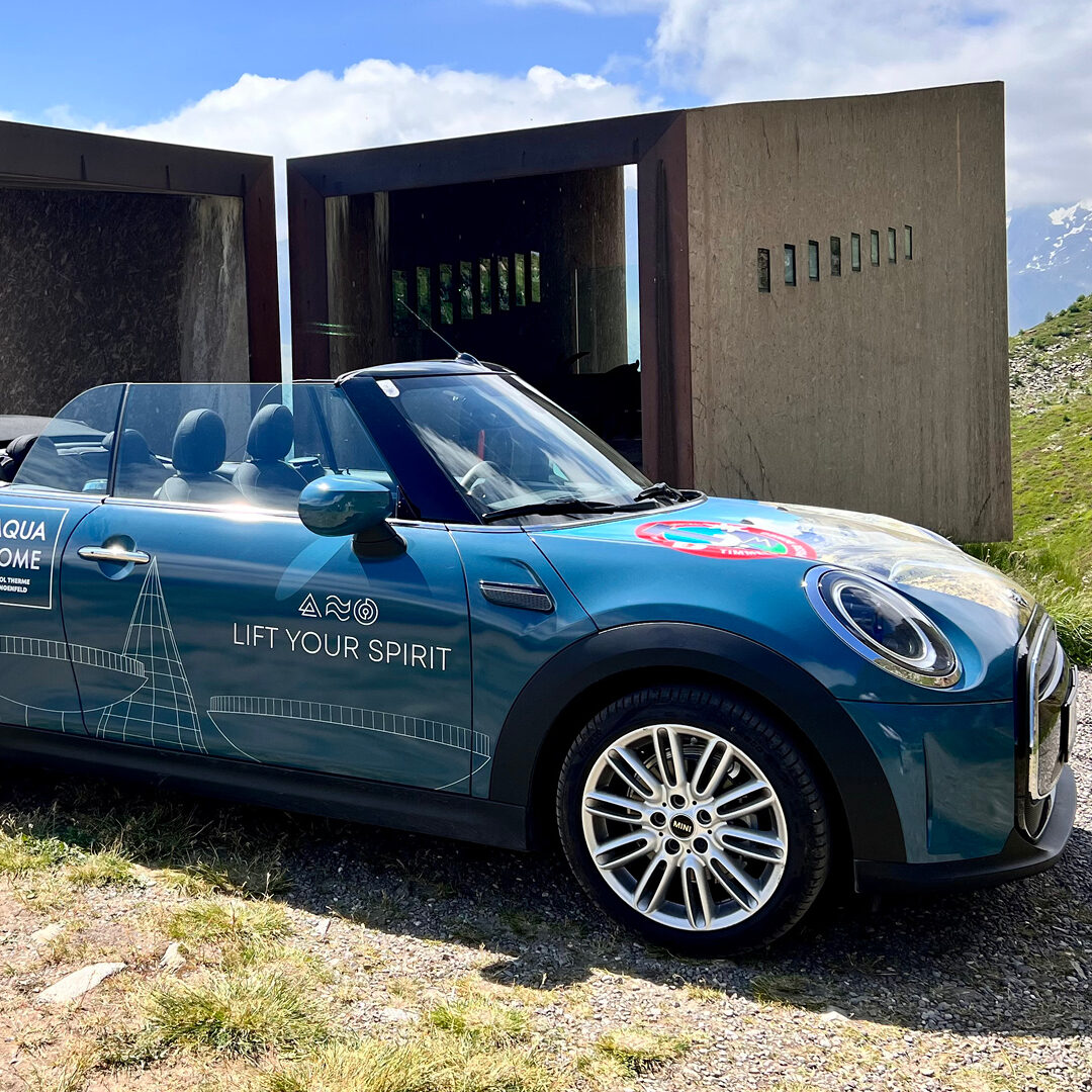 Voiture décapotable bleue avec des logos et les mots « LIFT YOUR SPIRIT » garée près de structures en béton modernes et d'un paysage herbeux avec des montagnes en arrière-plan.