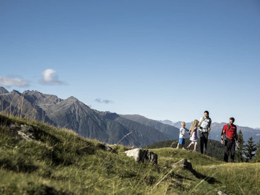 Группа из четырех человек, включая двух детей, идет по травянистой горной тропе на фоне далекой горной гряды под ясным голубым небом.