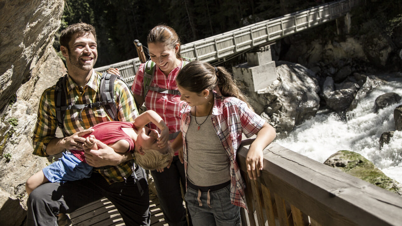Семья из четырех человек, одетая в верхнюю одежду, стоит на деревянном мосту у реки; отец игриво держит маленького ребенка вверх ногами.