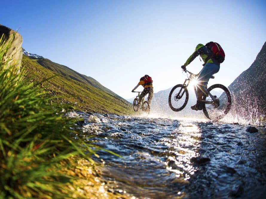 Два велосипедиста едут по мелкому ручью среди горного пейзажа под ясным голубым небом.