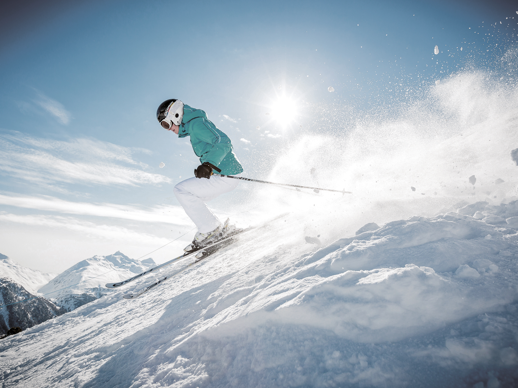 Person fährt unter klarem blauen Himmel, mit Gischt im Hintergrund und hellem Sonnenlicht einen schneebedeckten Hang hinunter. Trägt blaugrüne Jacke, weiße Hose, Helm und Schutzbrille.