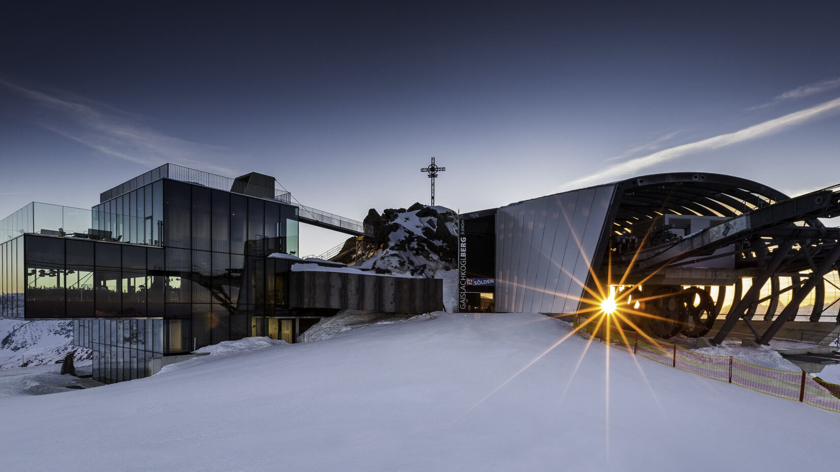 Modern mountain-top building with glass facade and mechanical structure, surrounded by snow at dawn. Sunlight beams through a large wheel, casting a starburst effect.