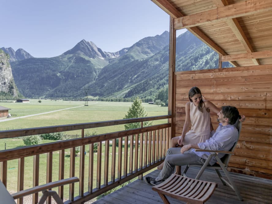 A couple relaxes on a wooden balcony with a scenic view of green fields and mountains under a clear blue sky, dreaming of their upcoming visit to the nearby thermal spa.