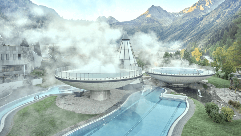 Steaming outdoor pools, shaped like large bowls, create a serene thermal spa experience against a breathtaking backdrop of mountains and trees.
