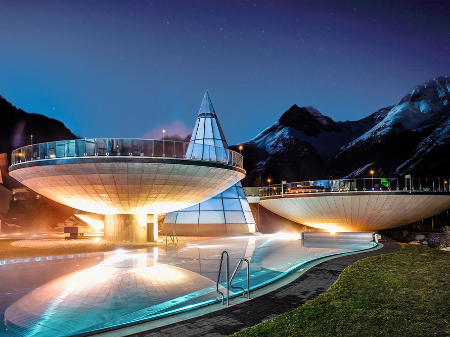 Futuristic, dome-shaped buildings with glass and metal structures illuminated at night, nestled around a steaming thermal spa pool, set against a backdrop of majestic mountains.