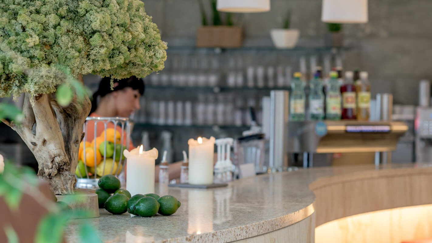 Eine gemütliche Café-Theke mit Kerzen, einer Topfpflanze, Limetten und verschiedenen Flaschen versprüht den Charme eines Thermenrestaurants. Im Hintergrund erscheint eine Person neben Regalen und einem verschwommenen Kassenbereich, was die einladende Atmosphäre verstärkt.