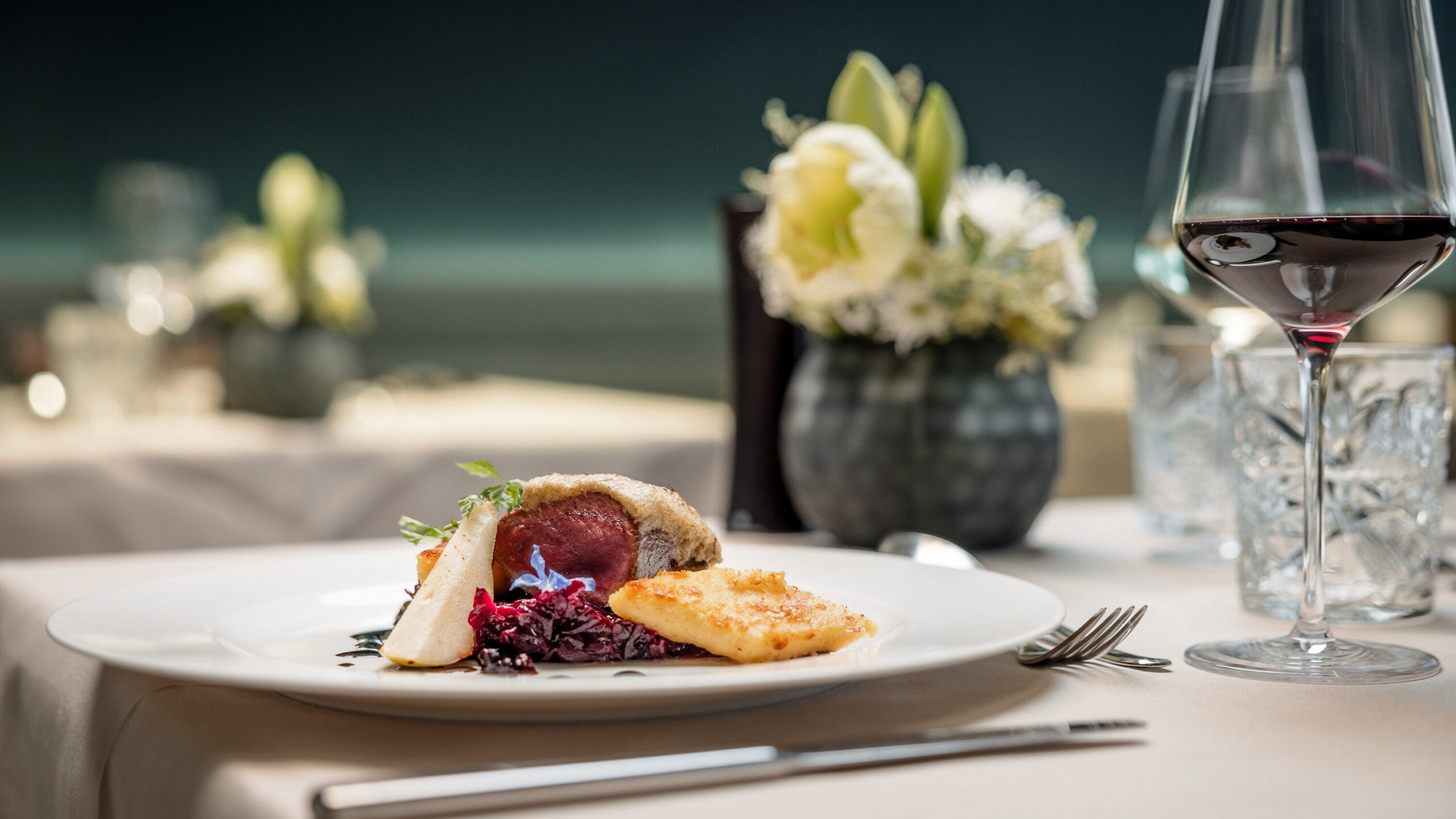 A gourmet meal featuring a succulent cut of meat, garnished greens, and a cubed side dish elegantly presented on a white plate. In the backdrop, a wine glass, flowers, and water glass adorn the dining table—a setting reminiscent of fine restaurants.