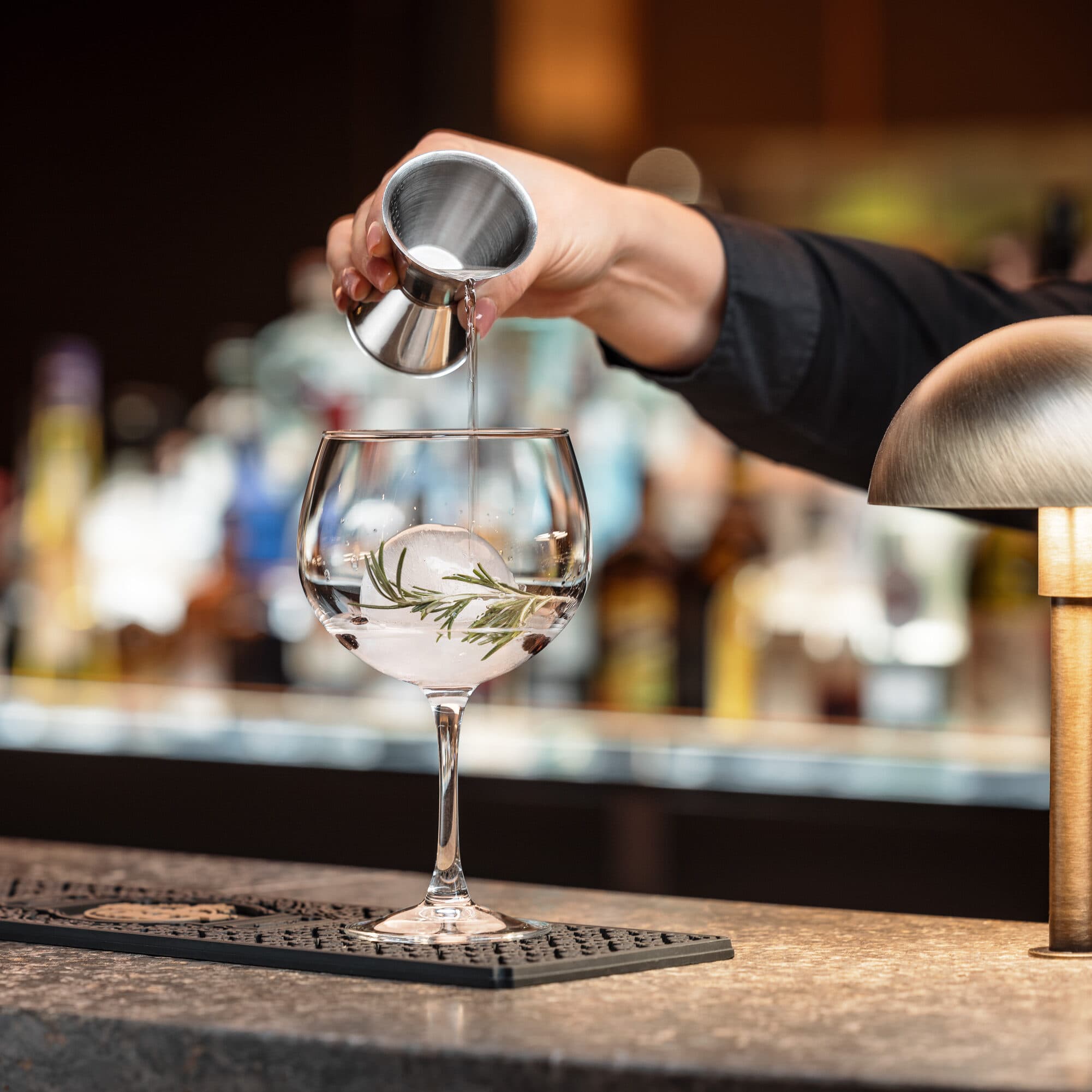 A bartender, expertly crafting cocktails in bustling restaurants, pours liquid from a jigger into a stemmed glass with ice and a rosemary garnish, all perfectly positioned on the bar counter.