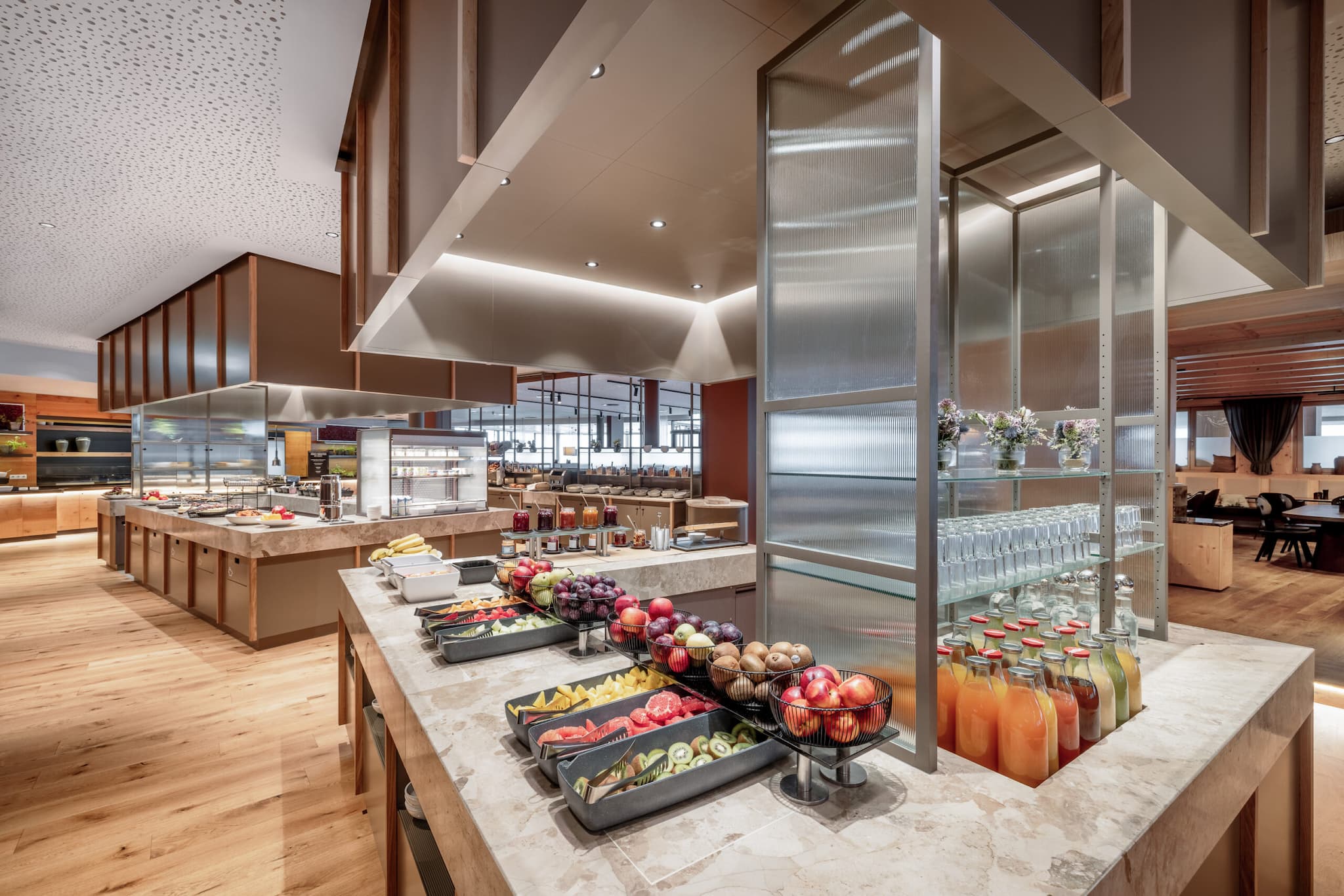 The spacious buffet area resembles top-tier restaurants, boasting a variety of fruits, juices, and glasses on display. The modern interior features wooden flooring and ceiling lights, creating an inviting ambiance.