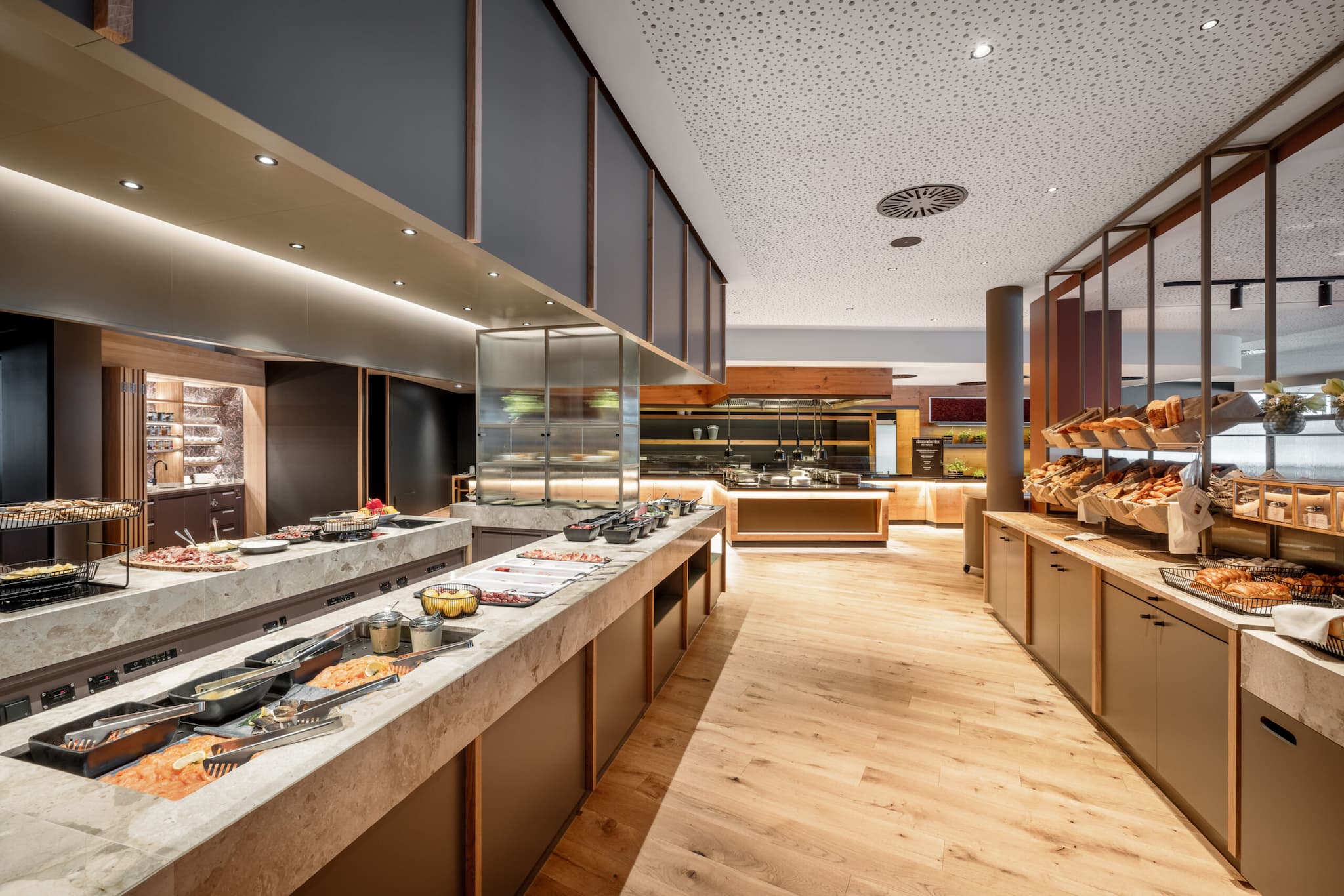 The modern buffet area, reminiscent of upscale restaurants, boasts marble countertops and wooden flooring, showcasing a variety of breads, pastries, and dishes. The ceiling features recessed lighting and perforated panels for an elegant dining ambiance.