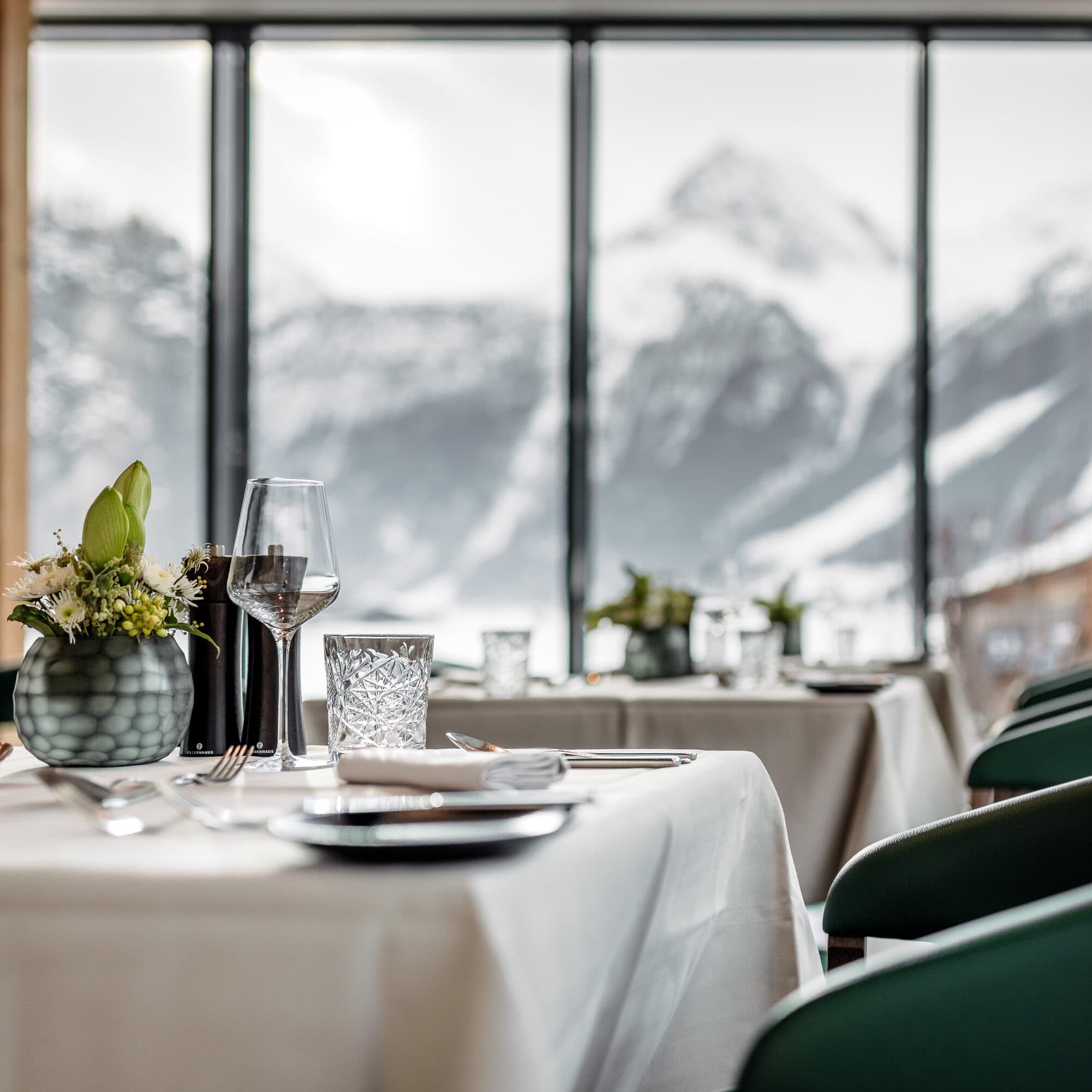 An elegant restaurant setting showcases wine glasses and flowers on tables by large windows, offering a stunning view of the snowy mountain landscape outside.
