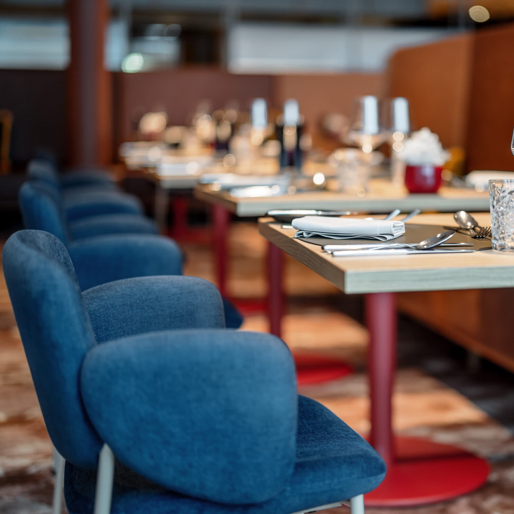 Empty restaurant tables with blue chairs are perfectly set in this inviting space, their wooden surfaces adorned with wine glasses, silverware, and napkins, embodying the elegance found in upscale restaurants.