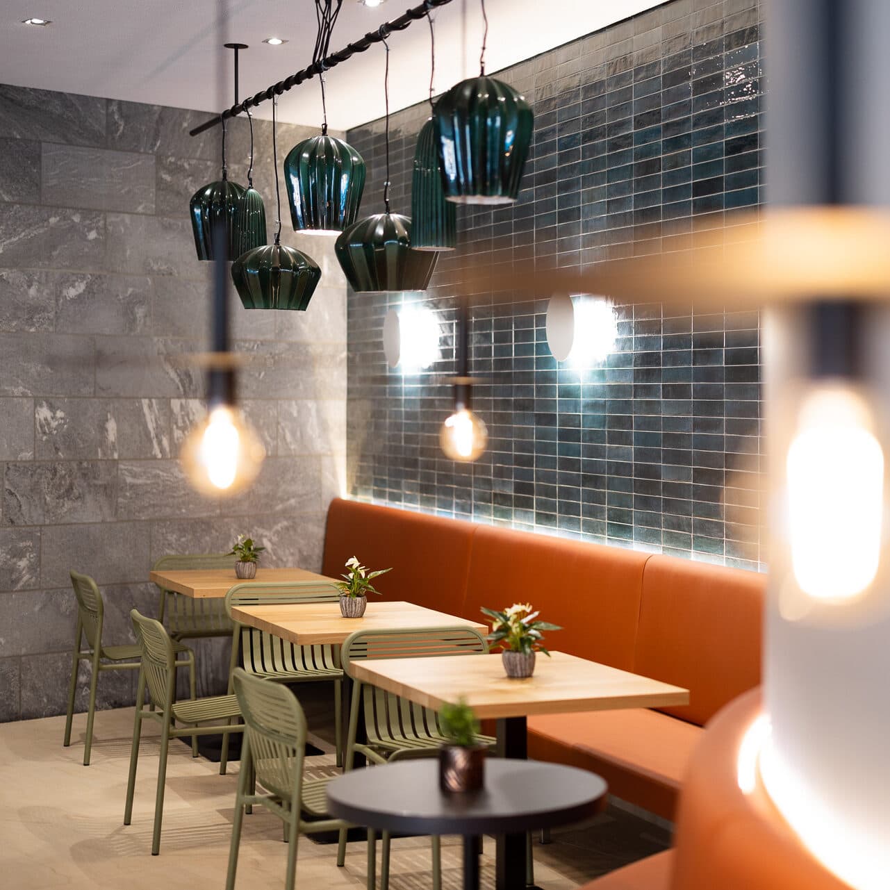 Modernes Café-Interieur im Stil eines Thermenrestaurants mit grünen Pendelleuchten, gemütlichen grünen Stühlen und hellen Holztischen. Eine orangefarbene Polsterbank säumt die Wand mit schicken blauen Fliesen. Kleine Pflanzen zieren jeden Tisch und verleihen dieser ruhigen Oase einen Hauch von Natur.