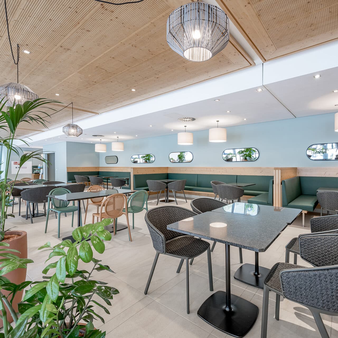 Ein modernes Thermenrestaurant mit Tischen, Stühlen und Deckenbeleuchtung. Grüne Pflanzen und hellblaue Wände mit kleinen Spiegeln werten das Dekor auf und schaffen eine ruhige Oase.