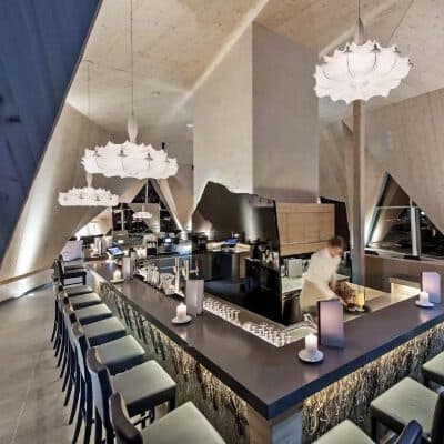 Moderne Bar in einem Thermenrestaurant mit eleganter Einrichtung, mit einer langen Theke, umgeben von Hockern, stilvollen Kronleuchtern und einem Barkeeper, der in einem zeitgenössischen Interieur mit schrägen Decken Getränke zubereitet.