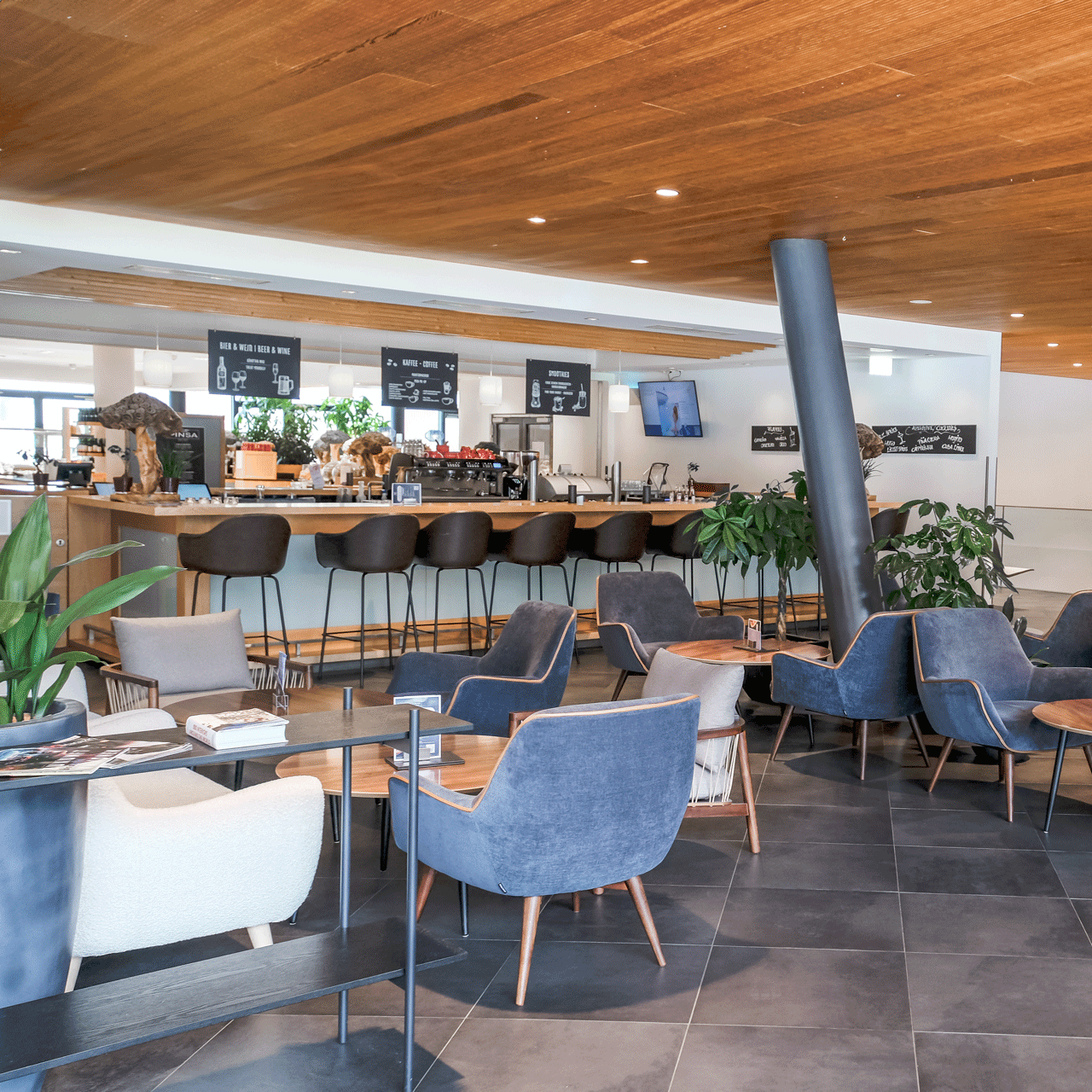 Das Thermenrestaurant besticht durch sein modernes Café-Interieur mit Holzdecke, blauen Polsterstühlen, Barhockern an der Theke, üppigen Grünpflanzen und gefliestem Boden. Hinter der Theke sorgen Menütafeln und ein TV-Bildschirm für eine einladende Atmosphäre.