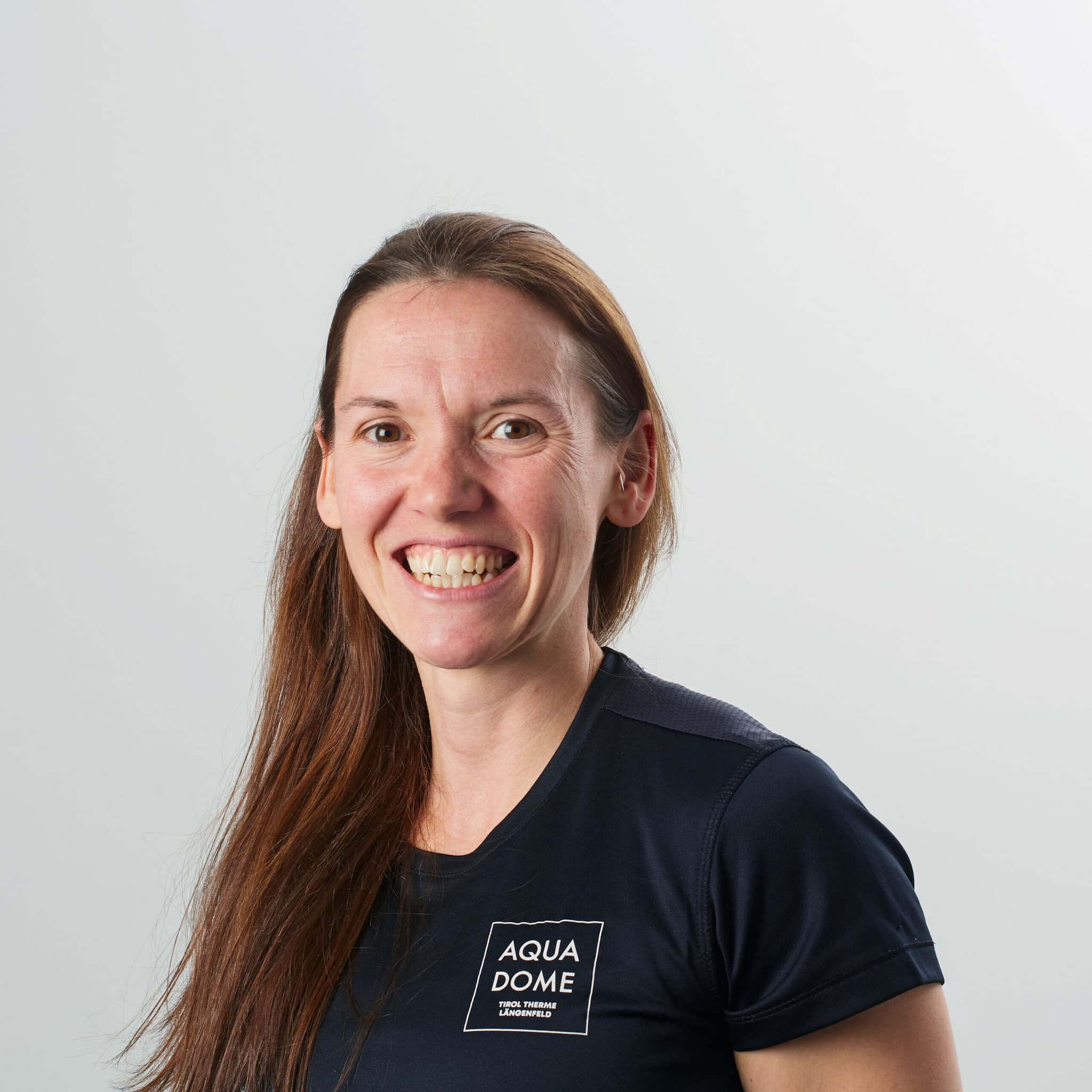 Frau mit langen braunen Haaren, die ein dunkles T-Shirt mit dem Aqua Dome-Logo trägt und vor einem hellgrauen Hintergrund lächelt.