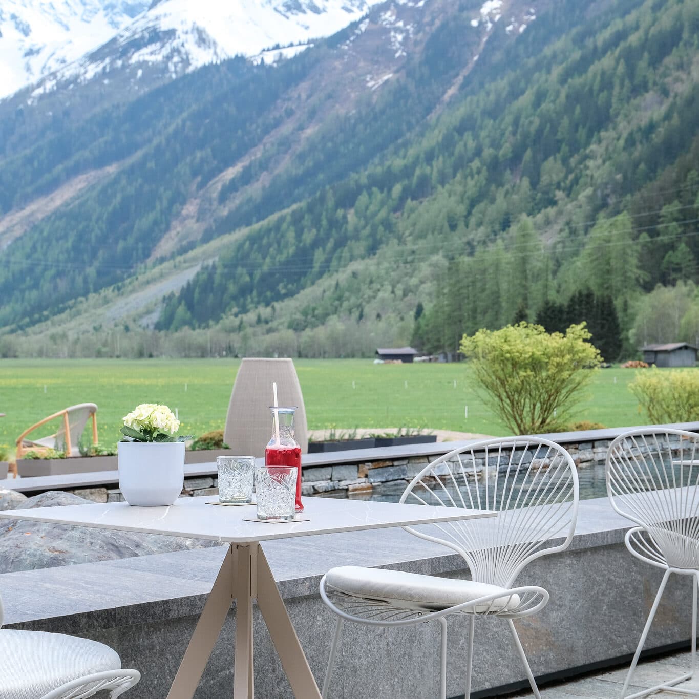 An outdoor patio at a restaurant featuring empty white wire chairs and a table adorned with drinks, all set against the stunning backdrop of mountains and forests.