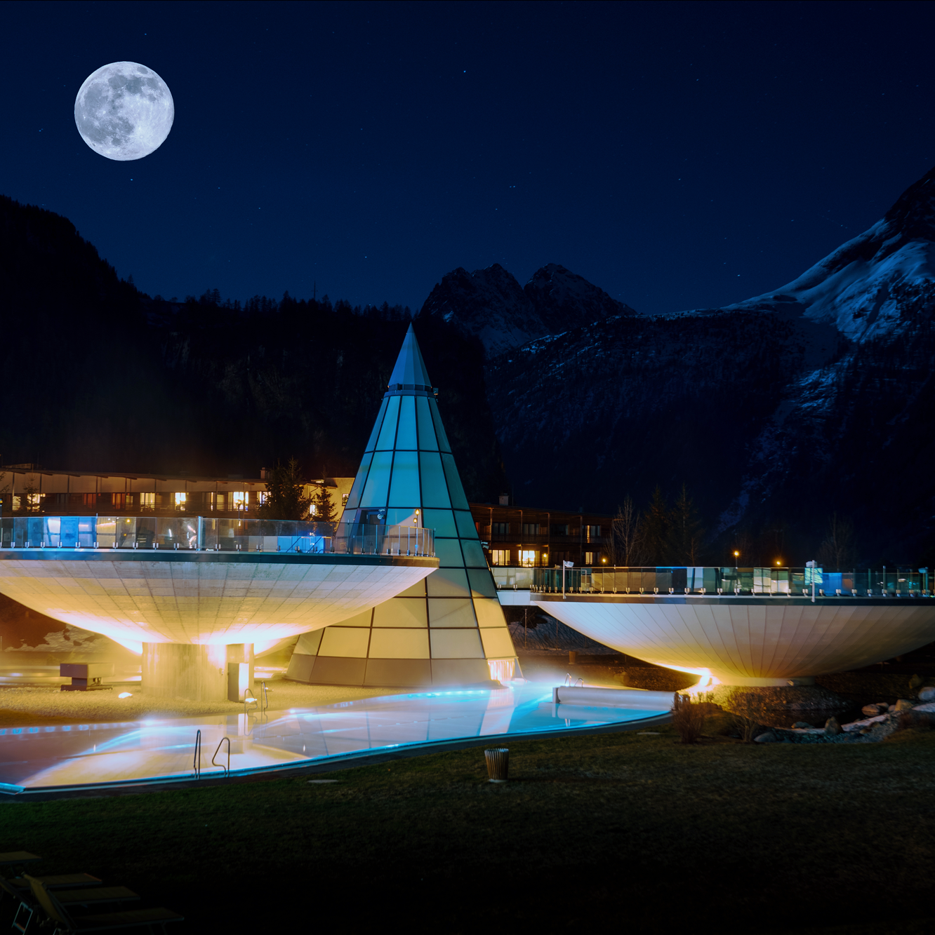 Modern, illuminated architectural structures with cone and bowl shapes stand against a mountainous backdrop under a full moon at night.