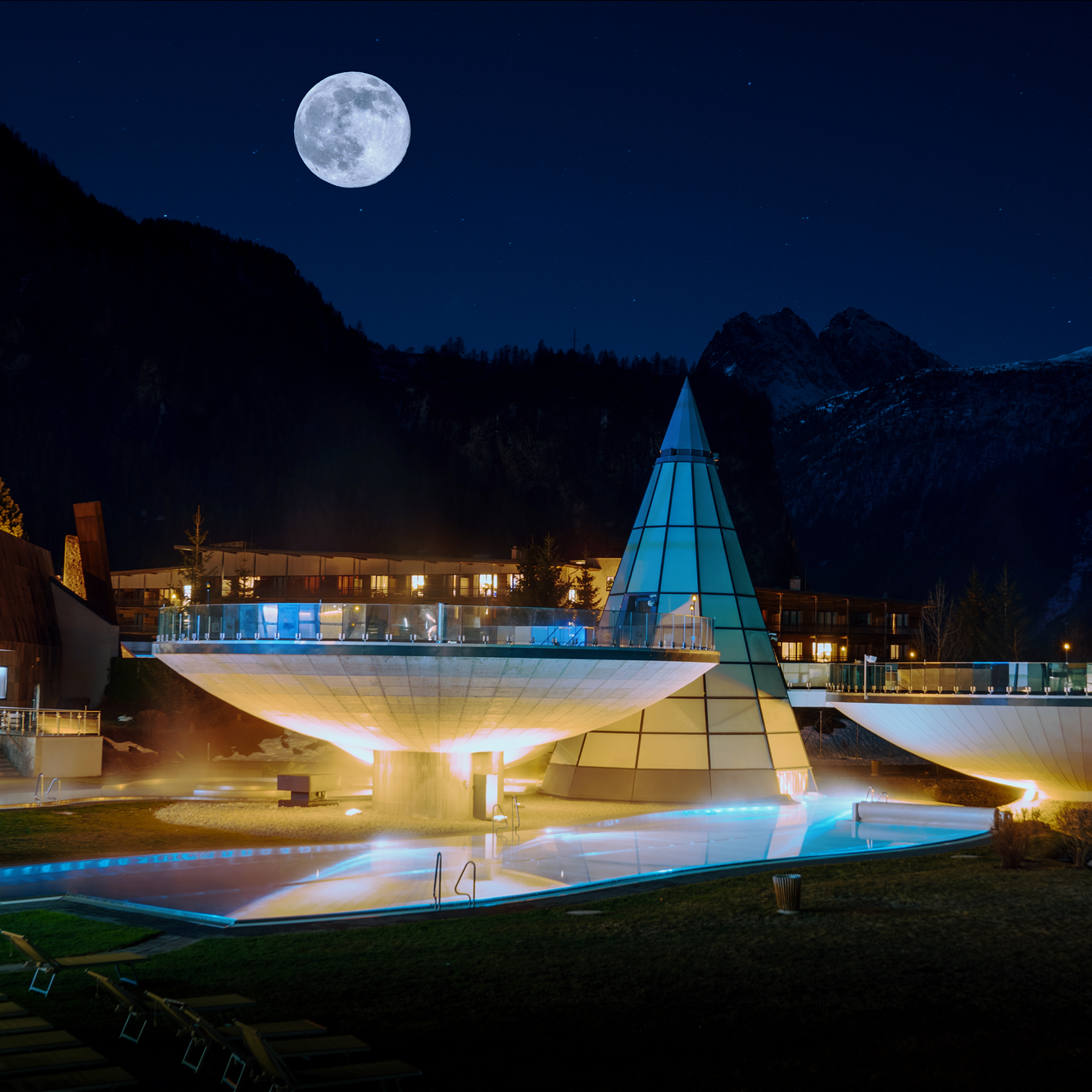 Nachts beleuchteter Außenpool des Resorts mit modernem architektonischem Design und einem Vollmond, der am klaren Himmel über den Bergen im Hintergrund sichtbar ist.