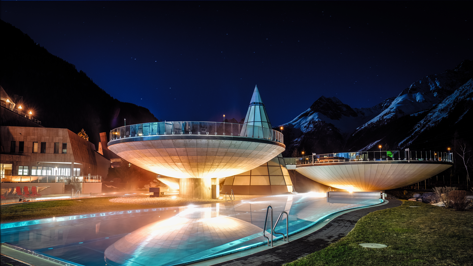 Modern architectural structures with conical roofs and glowing lights sit beside a reflective pool, surrounded by mountains under a starry night sky.
