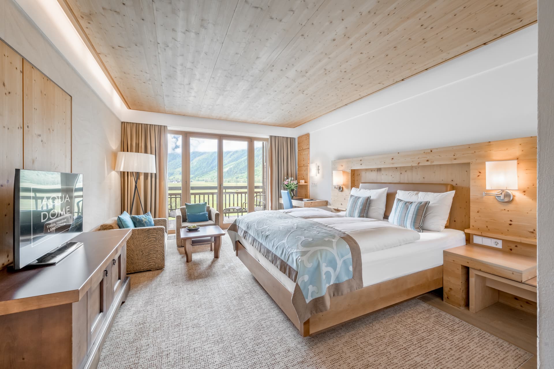 Modernes Hotelzimmer mit großem Bett, Sofa, Flachbildfernseher, Holzmöbeln und Balkontüren mit Blick auf die malerischen Berge.