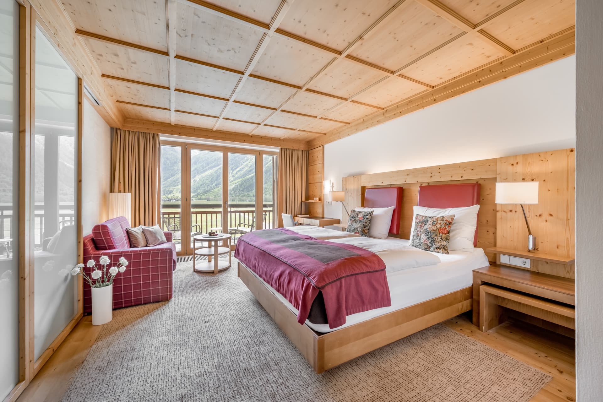 Современный гостиничный номер с двуспальной кроватью, диваном, небольшим столиком, деревянной мебелью, большими окнами и балконом с видом на горы.