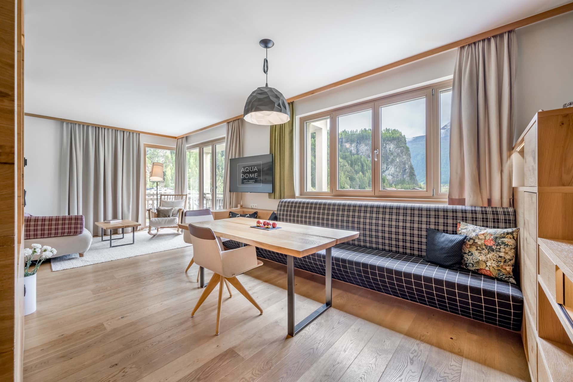 Светлая гостиная и столовая с деревянными полами, клетчатыми скамейками, деревянным столом, стульями, большими окнами с занавесками и видом на зелень и горы за окном.