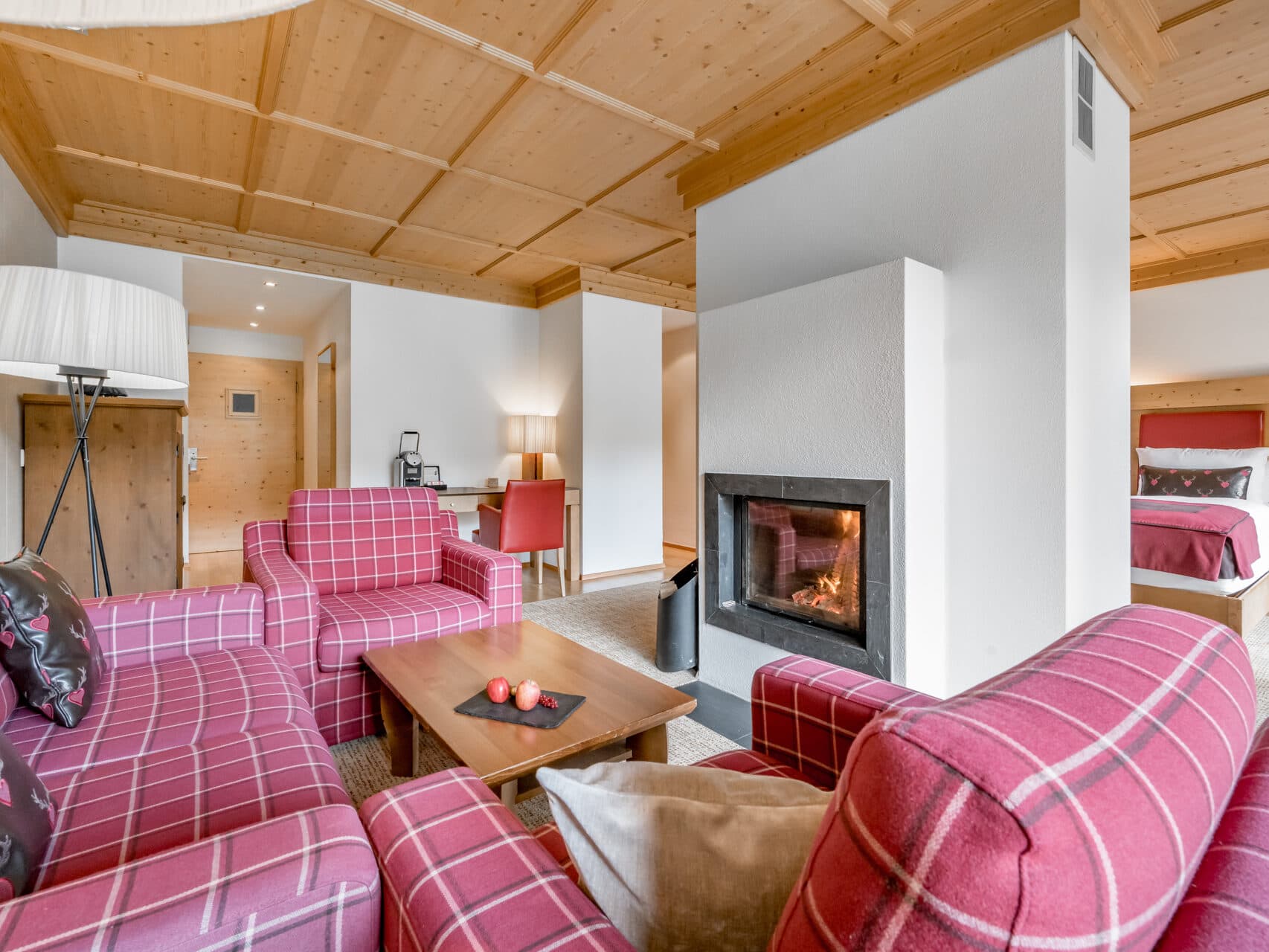 Eine gemütliche Hotelsuite mit rotkarierten Sesseln, einem hölzernen Couchtisch, einem Kamin, einem Bett im Hintergrund und holzgetäfelten Decken und Wänden.