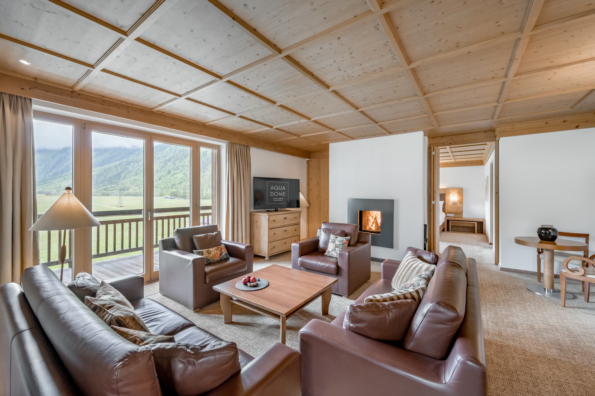 Modernes Wohnzimmer mit braunen Ledersofas, Holzdecke und -möbeln, großen Fenstern mit Blick auf die Berge, einem Fernseher und einem beleuchteten Kamin. Im Hintergrund ist der Essbereich zu sehen.