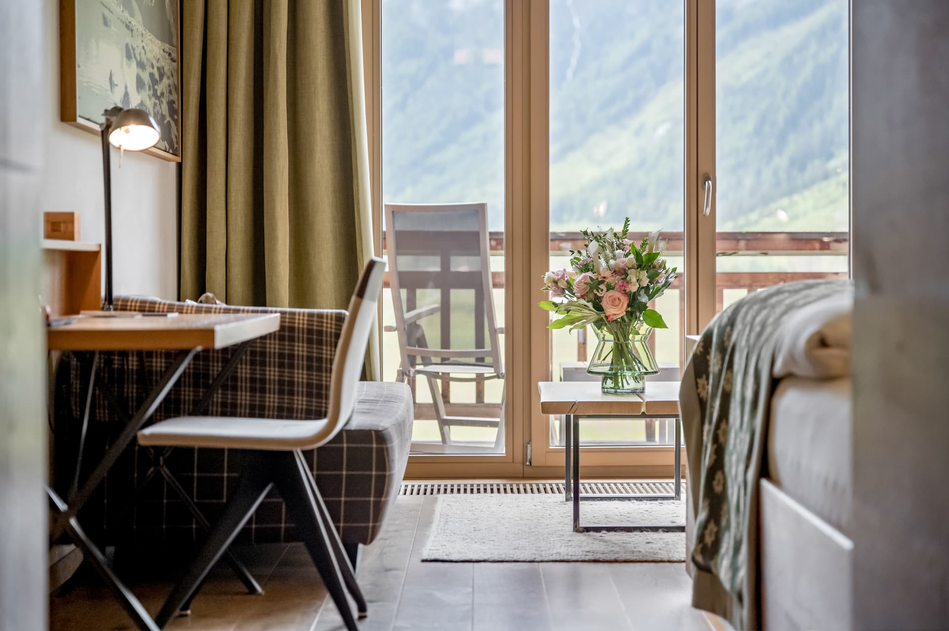 Ein Hotelzimmer mit Schreibtisch, Stuhl und Bett, mit einer Blumenvase auf dem Tisch und einem Balkon mit Bergblick durch große Glastüren.