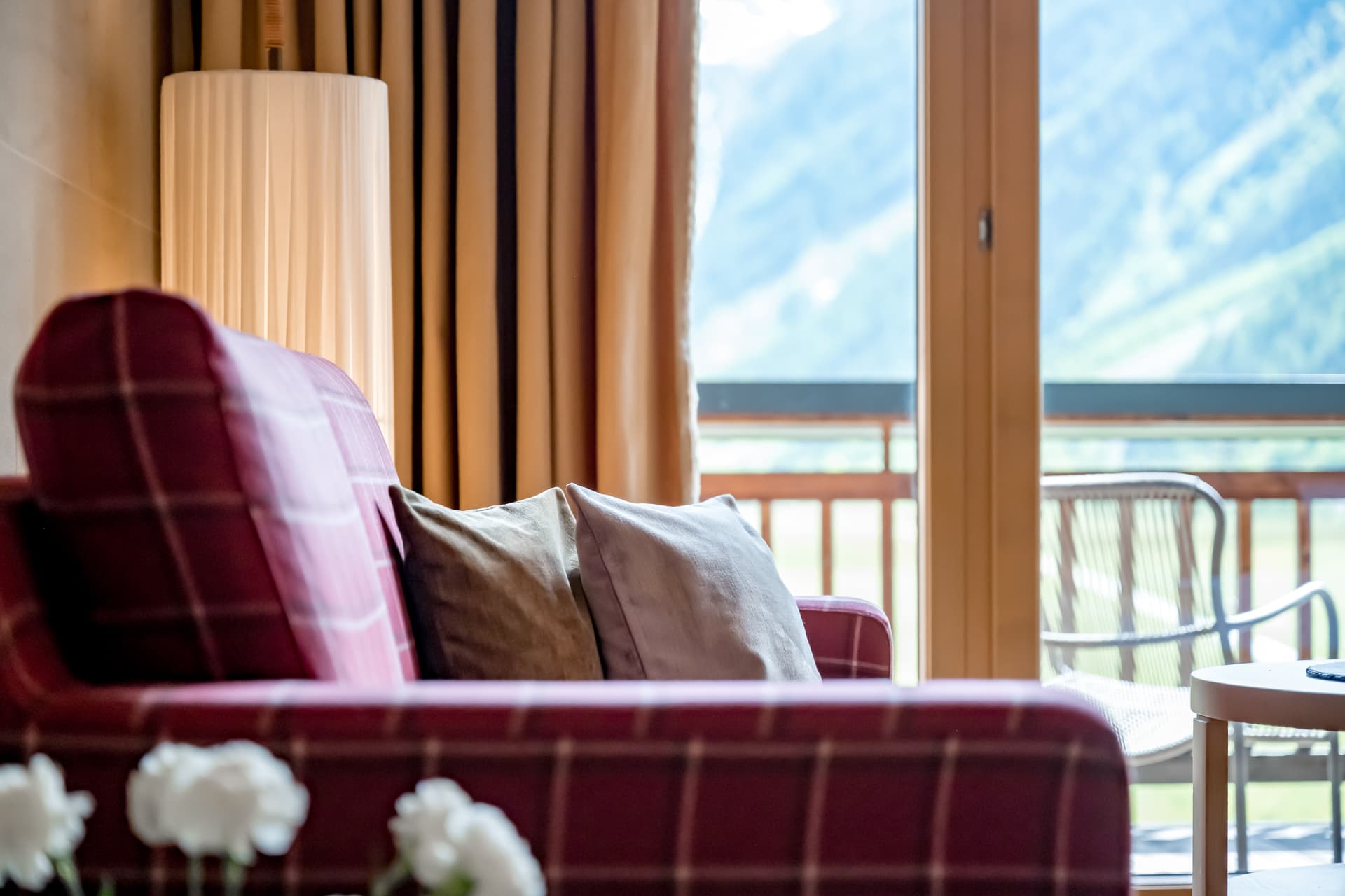 Красное клетчатое кресло с подушками стоит у окна, выходящего на балкон с видом на горы; здесь же можно увидеть торшер и кресло на открытом воздухе.