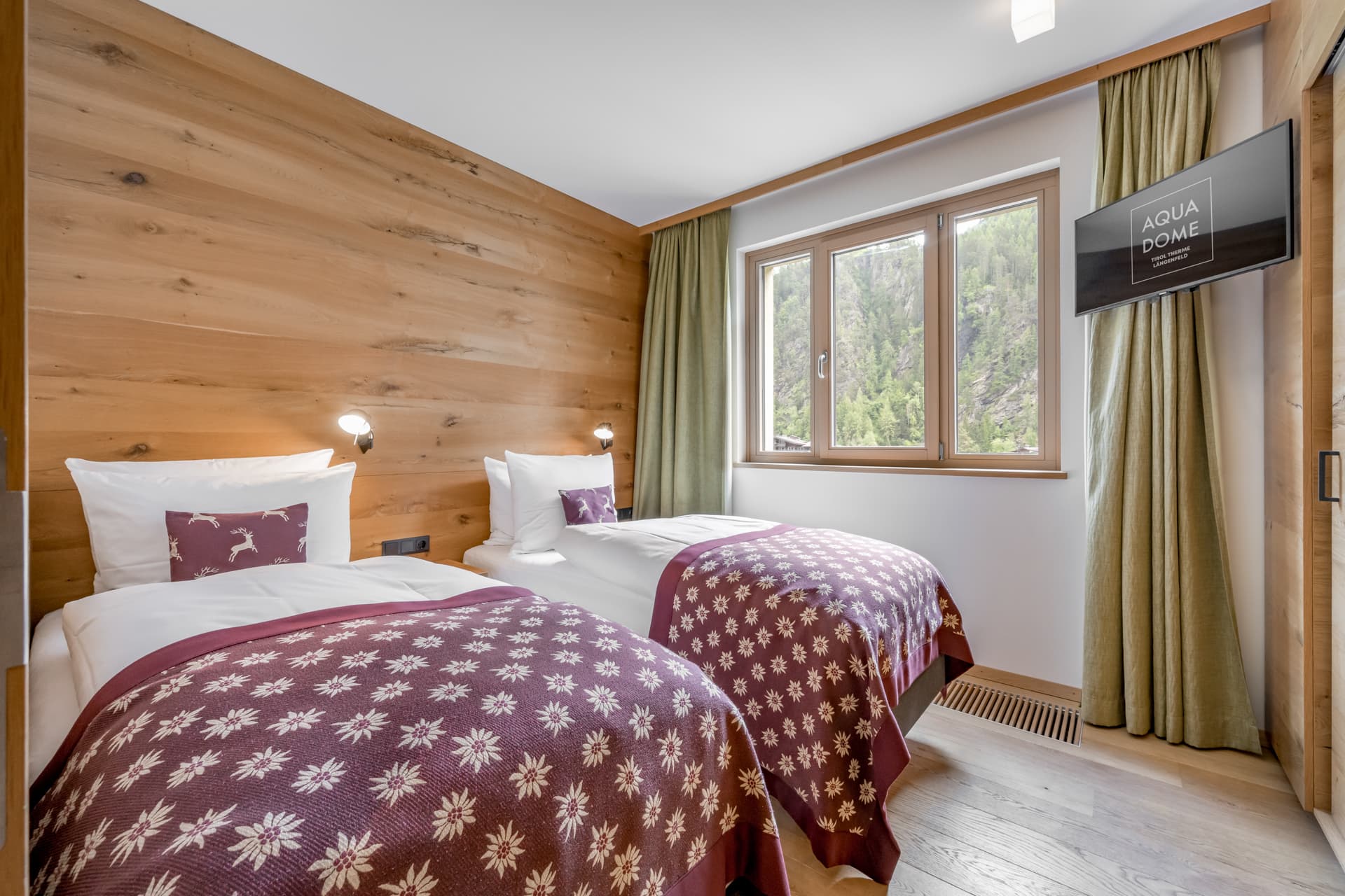 Гостиничный номер с двумя односпальными кроватями, покрывалами в цветочек, стенами, отделанными деревом, большим окном с занавесками и настенным телевизором с экраном AQUA DOME.