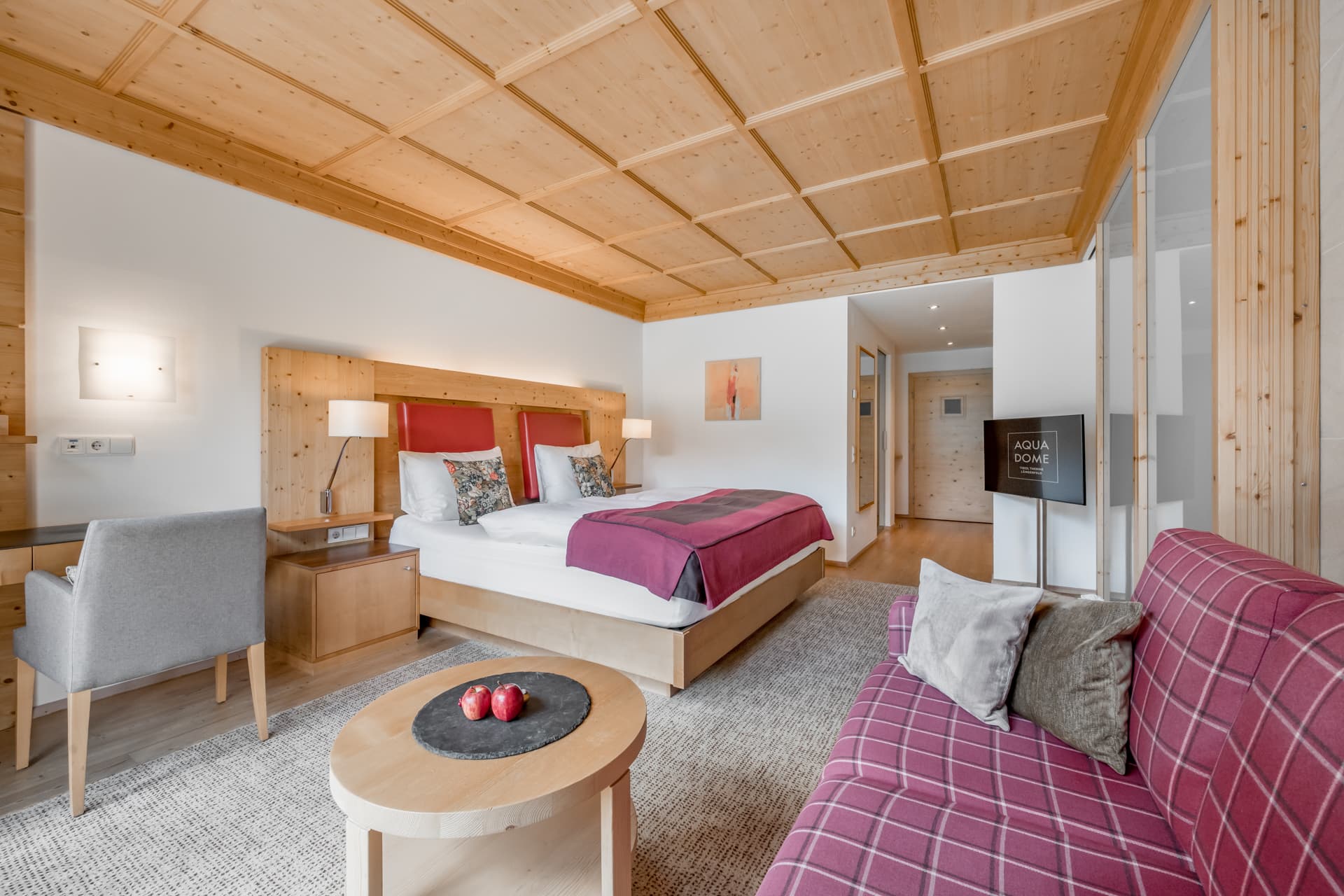 Современный гостиничный номер с деревянными акцентами, в котором есть двуспальная кровать с красными покрывалами, письменный стол, диван, круглый стол с яблоками и телевизор с плоским экраном.