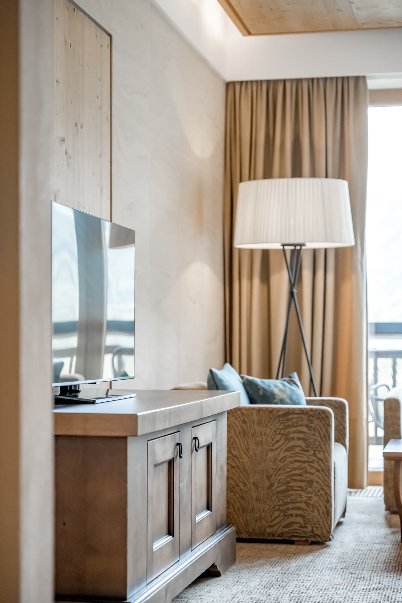 Ein Wohnzimmer mit einem hölzernen TV-Ständer, einem Flachbildfernseher, einem Sessel, einer Stehlampe und beigen Vorhängen an einem Fenster.