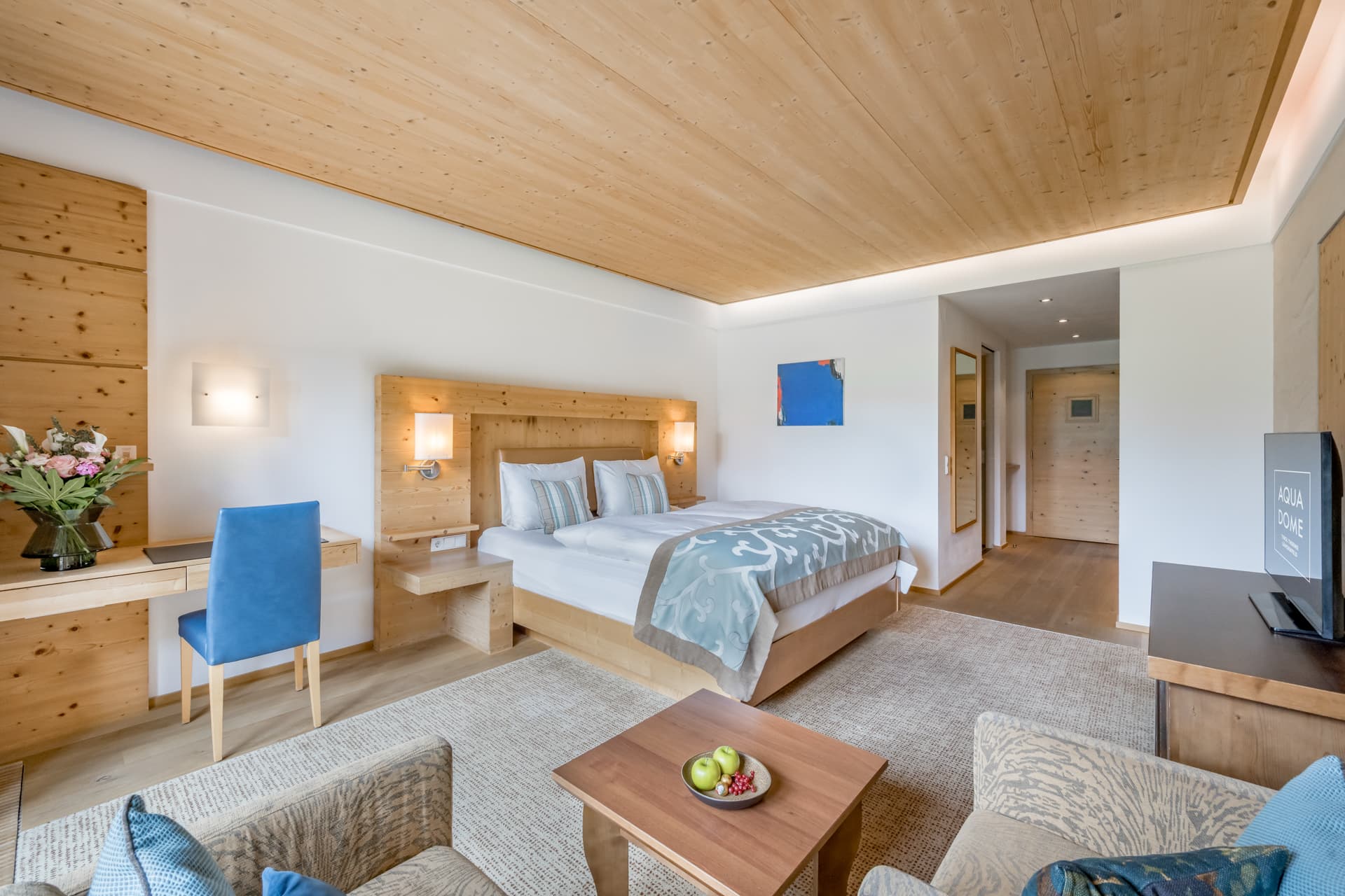 Modernes Hotelzimmer mit einem großen Bett, Holzmöbeln, einem Schreibtisch mit blauem Stuhl, einem Fernseher, einem Sofa und einem Couchtisch mit grünen Äpfeln. Neutrale Töne und natürliches Licht.