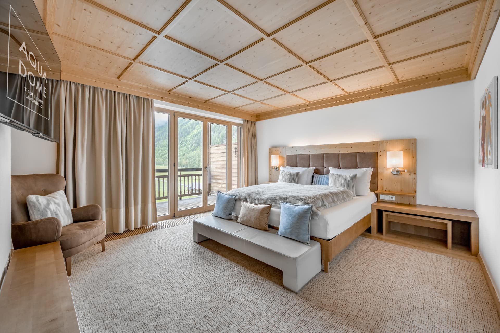 Modernes Hotelzimmer mit einem großen Bett, beigem Sessel, Holzdecke und bodenlangen Vorhängen. Eine Glastür führt auf einen Balkon mit Blick auf die Berge.